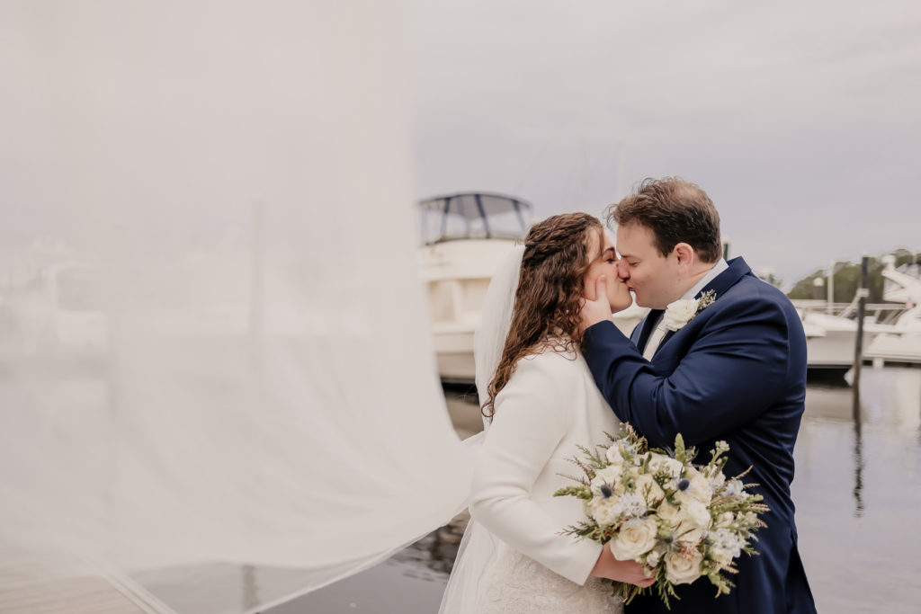 Wedding Couple kissing at a marina with yachts in the background. The bride's veil is flowing and stretched out.